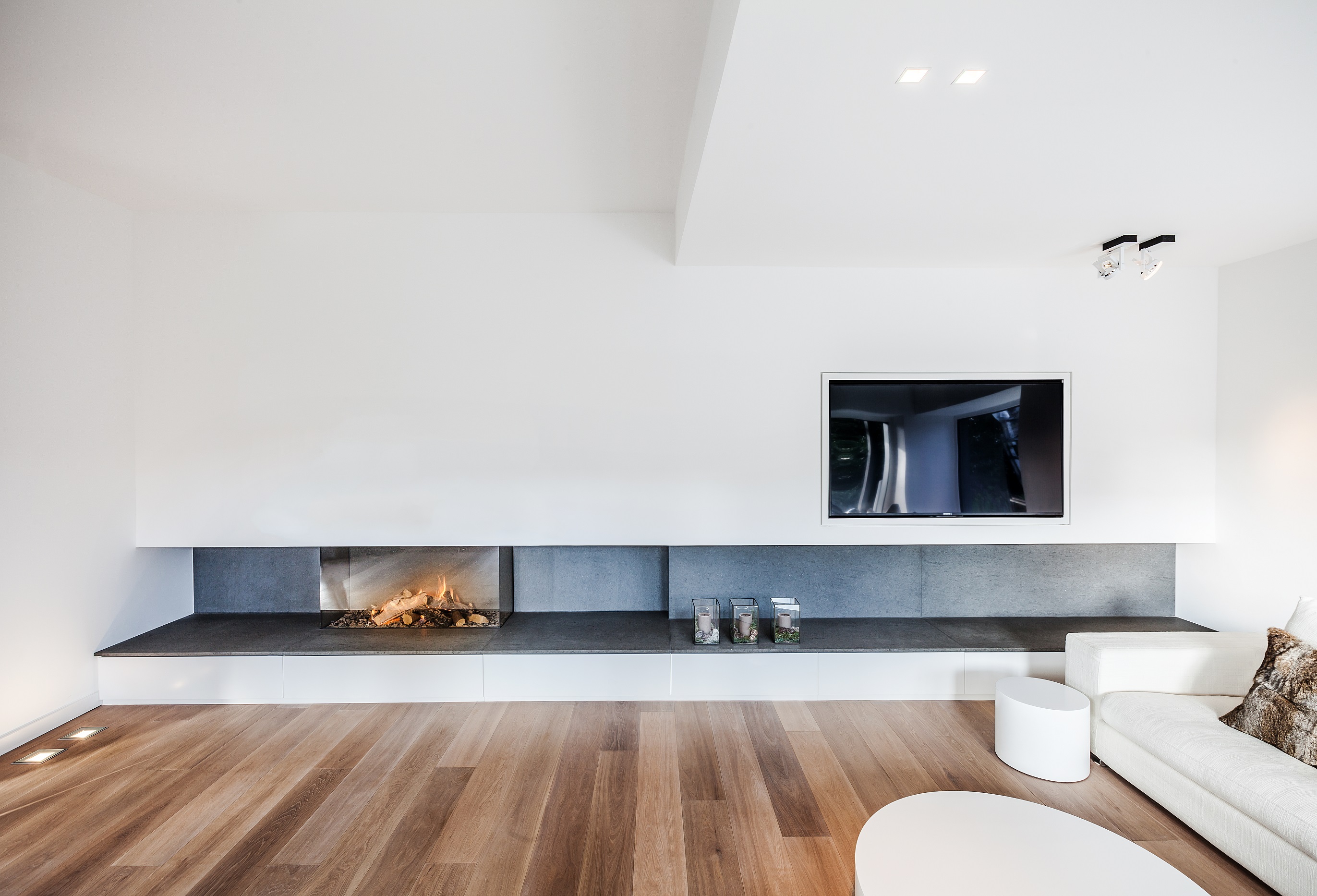 Zu sehen ist ein modern eingerichtetes Wohnzimmer mit Couch, Fernsehen, weißen Wänden, Kamin und Holz-Laminat.