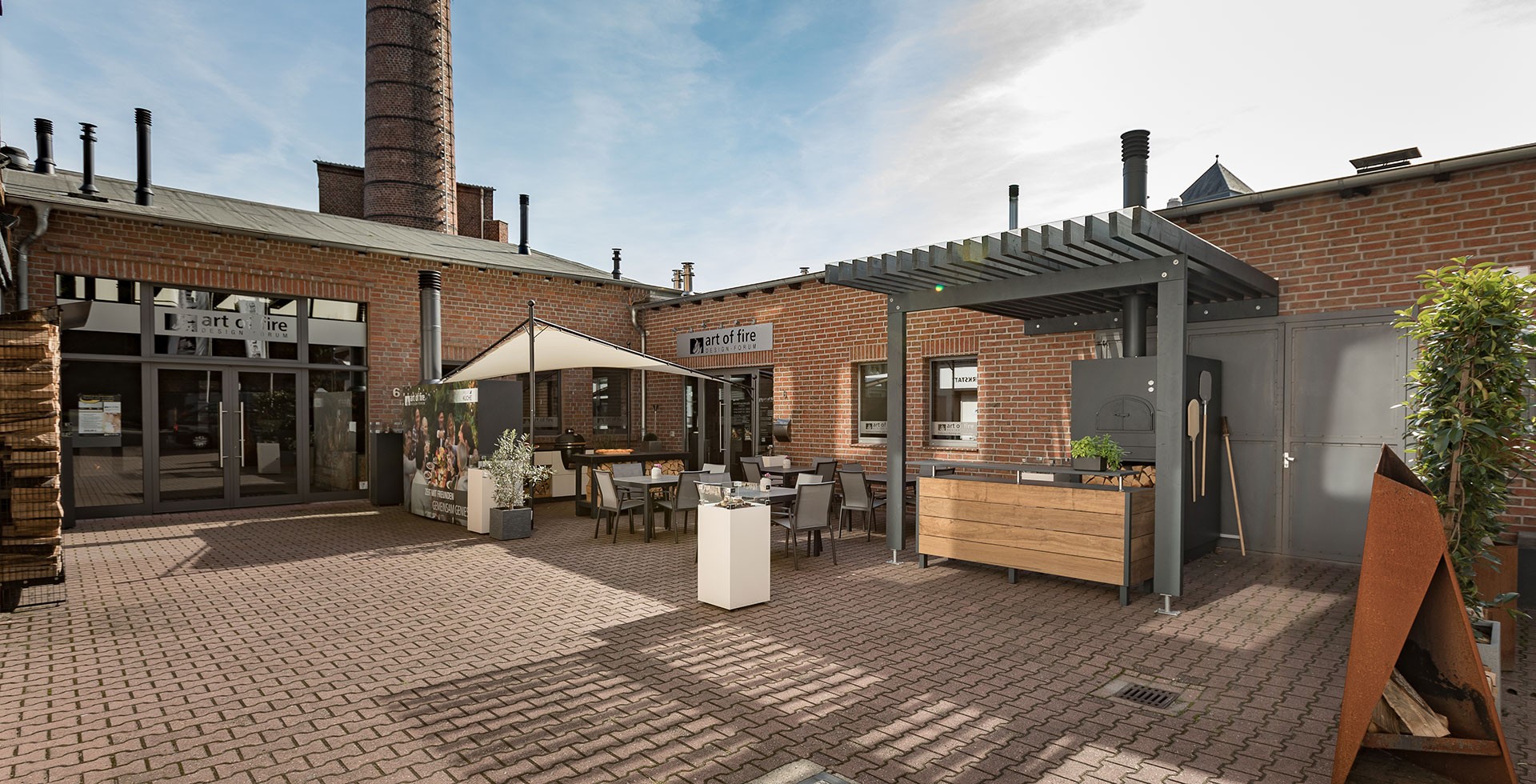 Zu sehen ist der Eingangsbereich der Filiale von art of fire. Es hat einen schönen außenbereich mit Sitzen, Sonnenschirm und großen Steinofen-Grill.