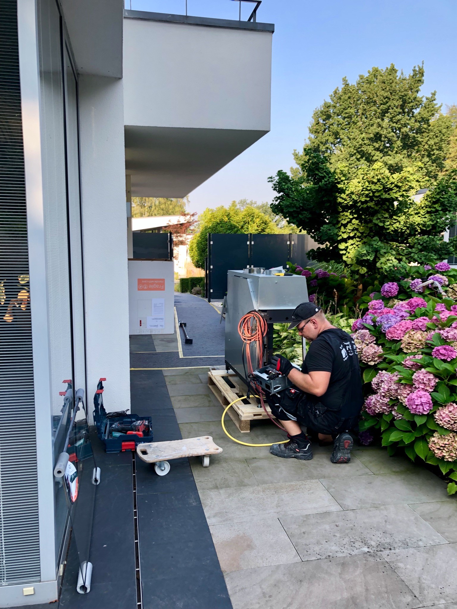 Zu sehen ist ein Handwerker welcher eine Kaminanlage im Vorgarten neben blühenden Hortensien montiert.