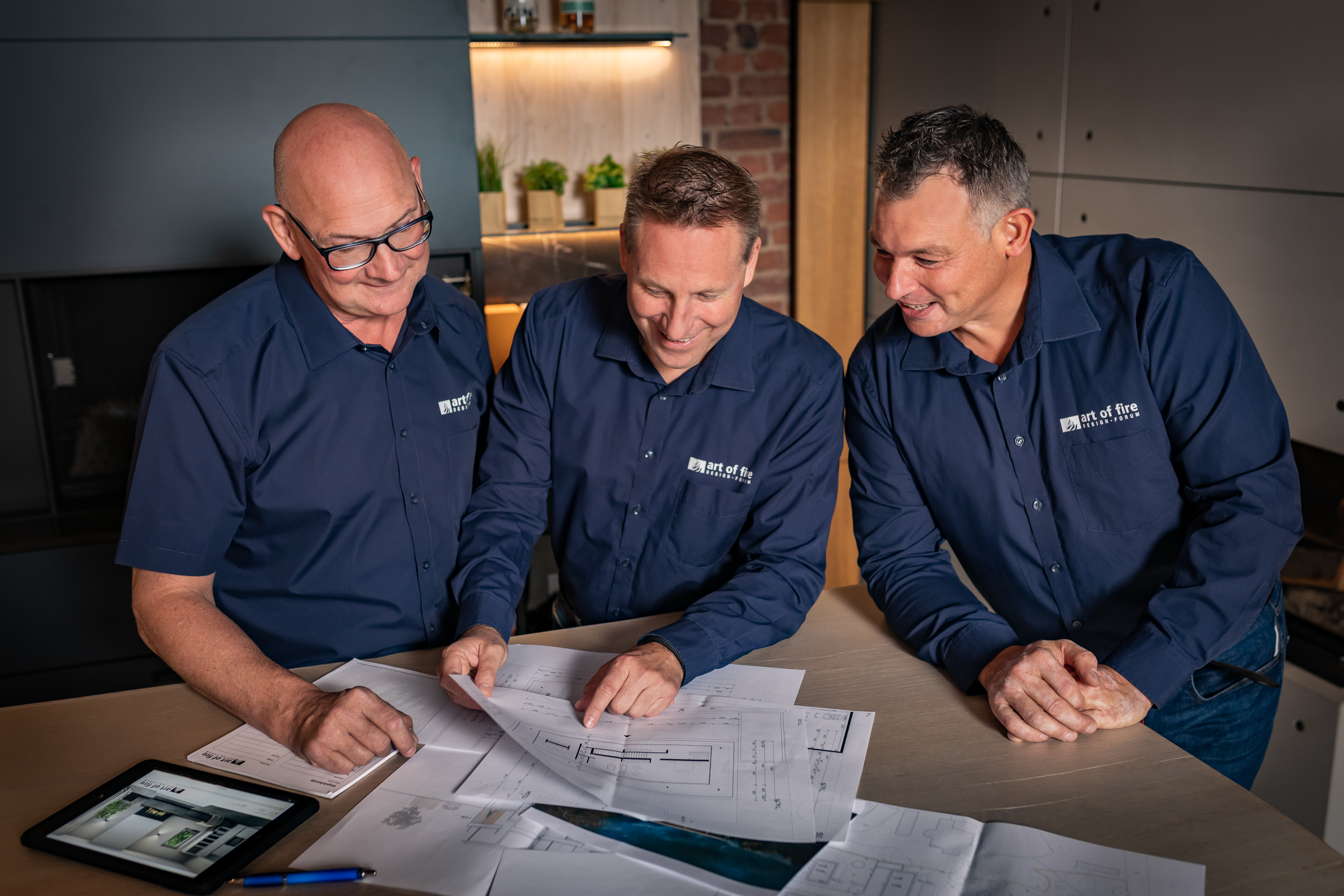 Zu sehen sind Drei Männer im Firmenhemd welche Baupläne an einem Tisch besprechen.
